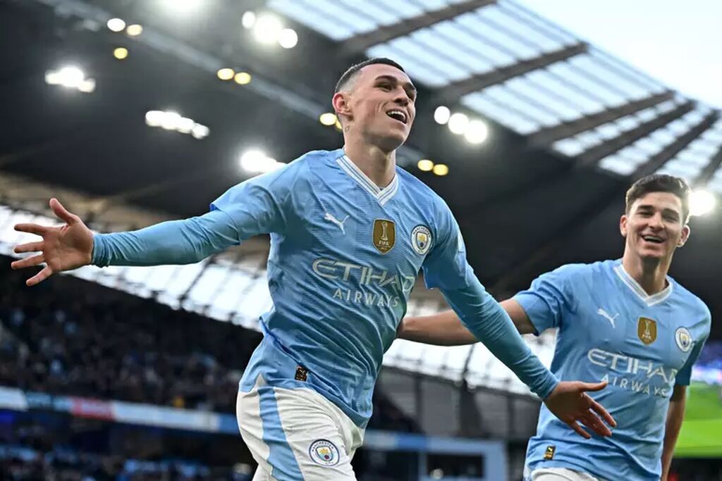 Jogador do Manchester City correndo com os braços abertos comemorando gol marcado durante partida.