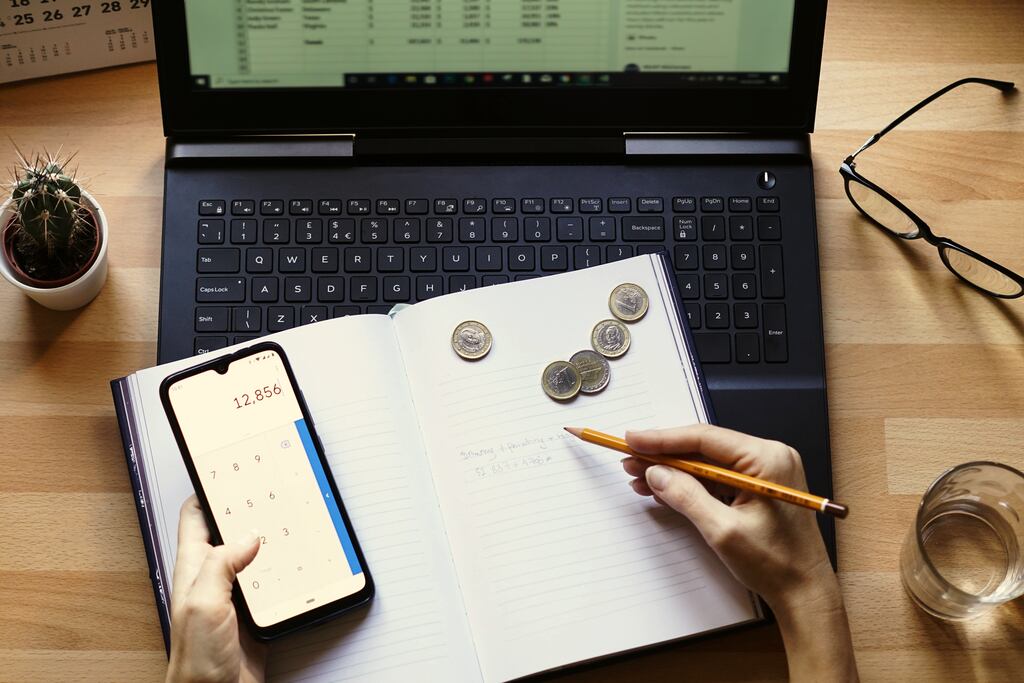 Pessoa segurando um celular com a calculadora aberta e escrevendo com outra mão em frente a um computador