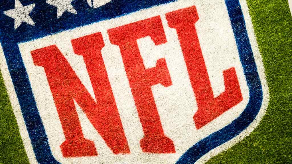 Escudo da NFL com suas cores branco, azul, e vermelho, desenhado em um gramado verde.