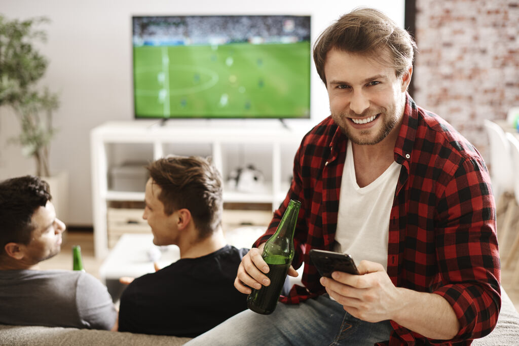 Homem de blusa branca e casaco quadriculado com vermeho e preto, tomando cerveja enquanto passa o jogo na TV ao fundo.