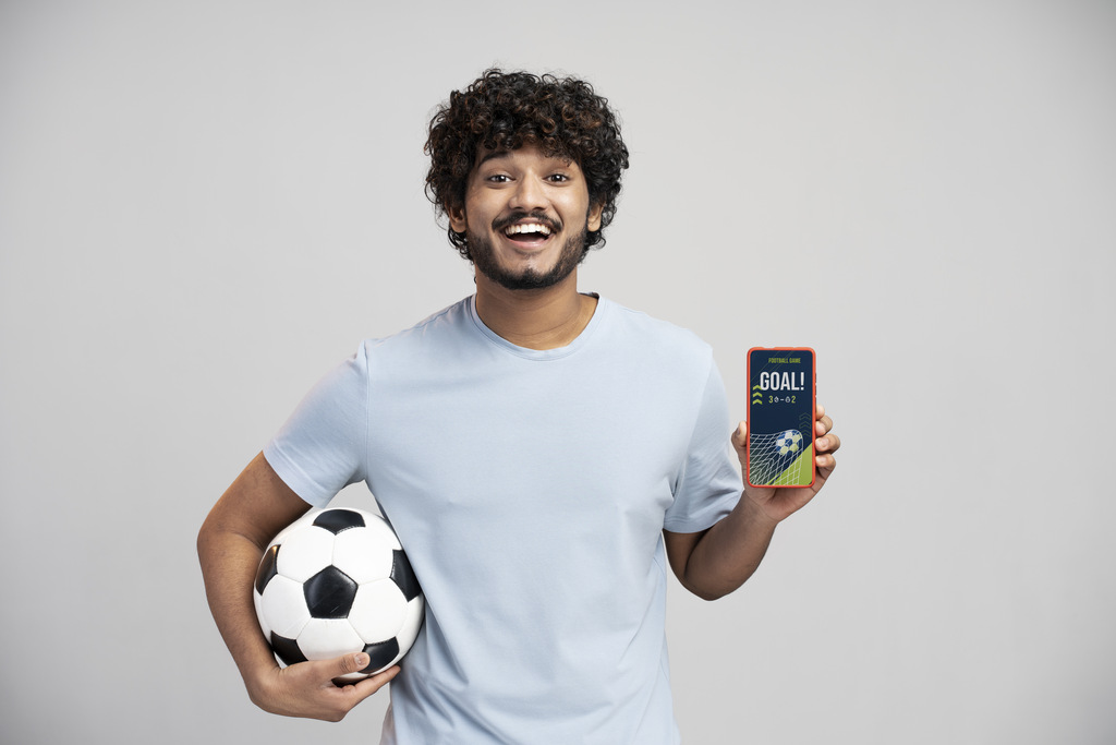 Homem de camisa azul clara, segurando uma bola de futebol com o braço direito e o celular escrito "gol" com a mão esquerda.