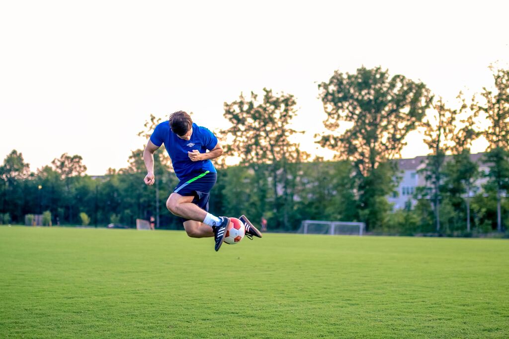 Jogador de uniforme azul, em campo de futebol no fim da tarde, dando do drible "lambreta".