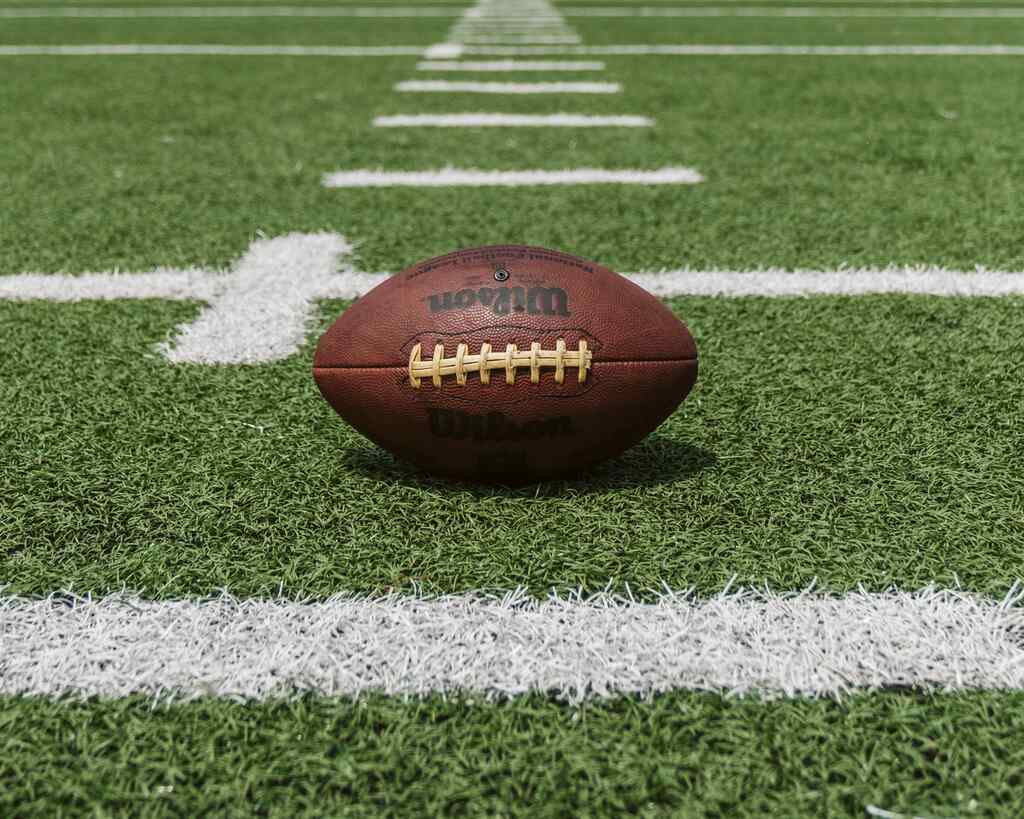 Bola de futebol americano colocada sobre a a grama e entre as linhas brancas de um campo de futebol americano.