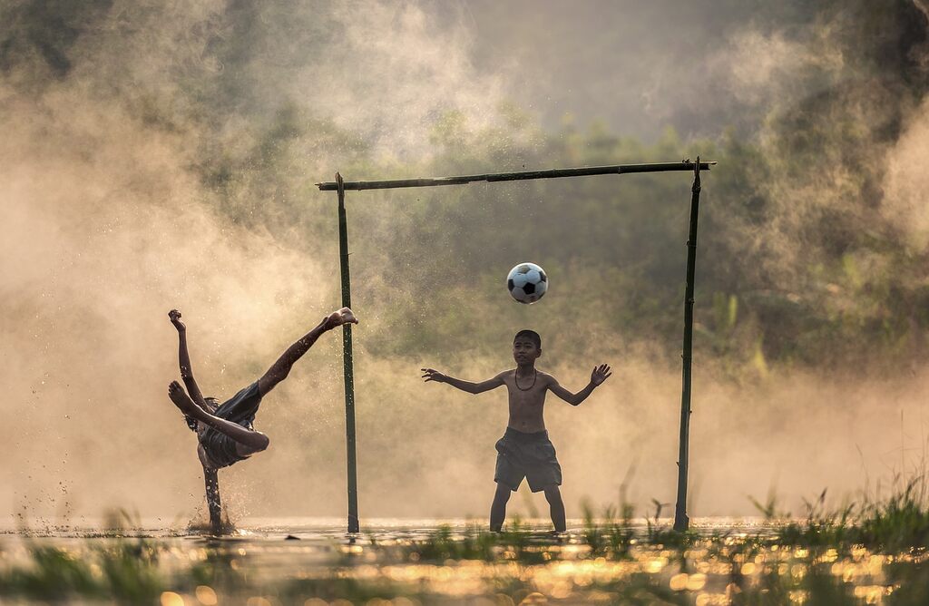 Menino dando bicicleta e bola indo em direção ao gol feito de madeira para outra criança defender.