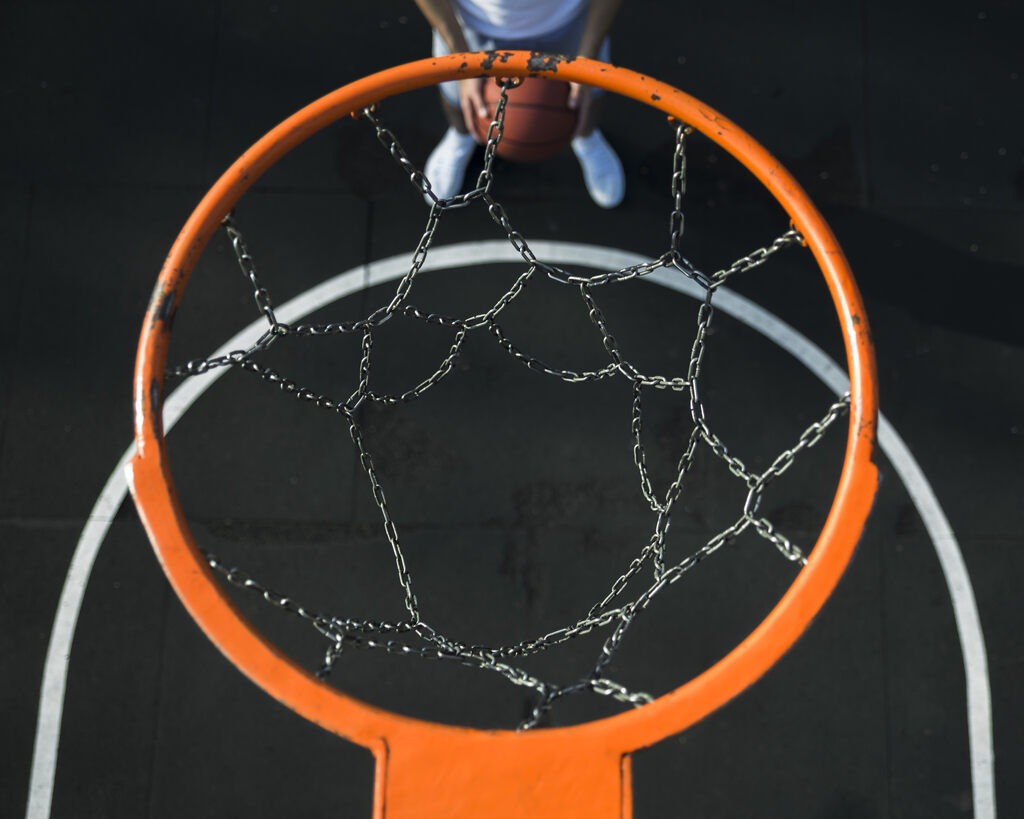 Cesta de basquete com aro laranja, rede escura e quadra preta com linha branca. Jogador segura bola de basquete.