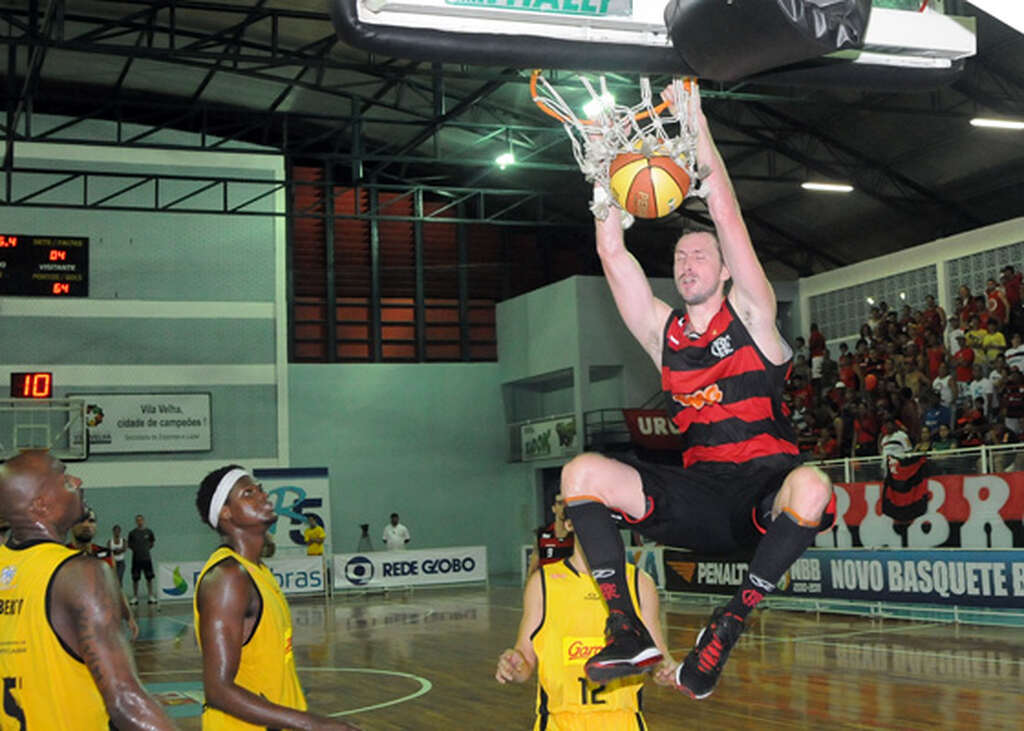Jogador do time de basquete do Flamengo realizando uma enterrada durante a partida, enquanto jogador adversário observa.