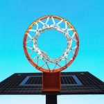 Cesta de basquete, com aro laranja e rede branca, vista bem debaixo da mesma com o céu azul de fundo.