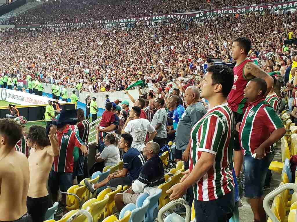 Torcida do Fluminense lotando o estádio, com muitos torcedores em pé, usando as cores do clube durante partida.