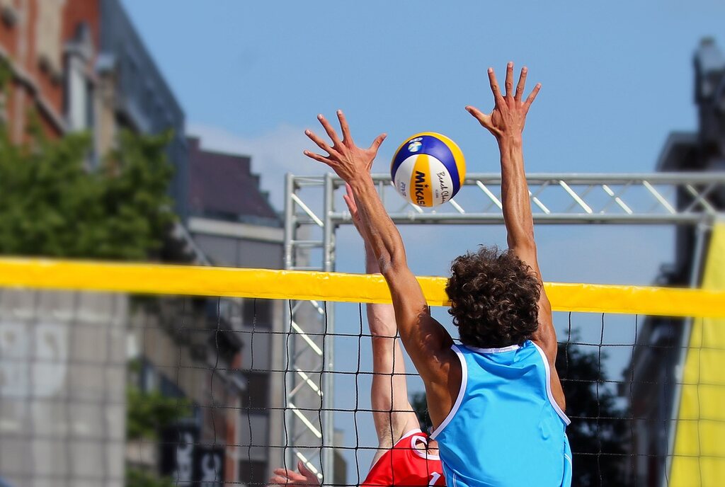 Jogador de vôlei de camisa regata azul-claro tentando bloquear cortada do jogador de vermelho.