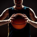 Jogador de basquete de blusa regata azul marinho, segurando a bola de basquete com as duas mãos na altura do peito.