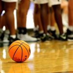 Bola de basquete laranja com linhas pretas, sobre um piso de madeira de uma quadra, com pernas de muitos jogadores ao fundo.