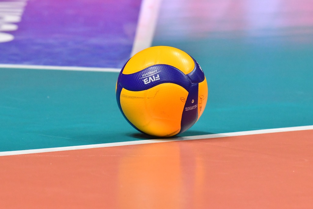 Bola de vôlei amarela, com detalhes azuis, parada sobre uma quadra de diversas cores.