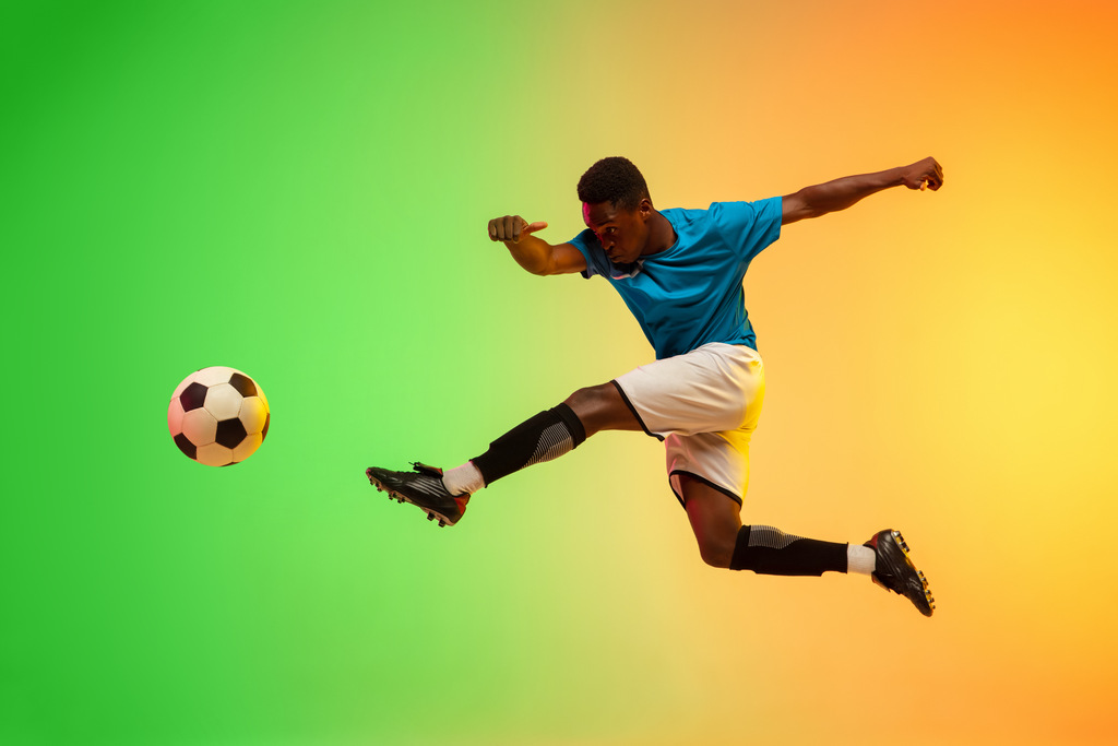 Jogador de futebol, negro, de uniforme azul, chutando uma bola com um fundo colorido atrás.