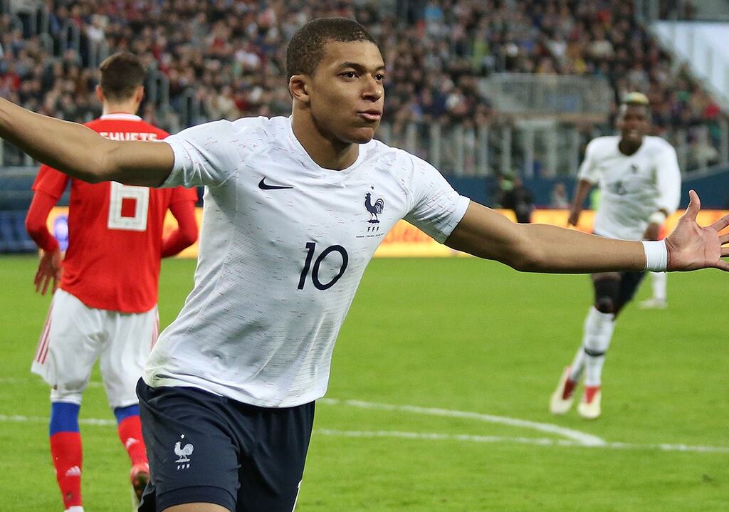 Kylian Mbappe comemorando gol de braços abertos com a camisa branca da seleção francesa durante partida de futebol.