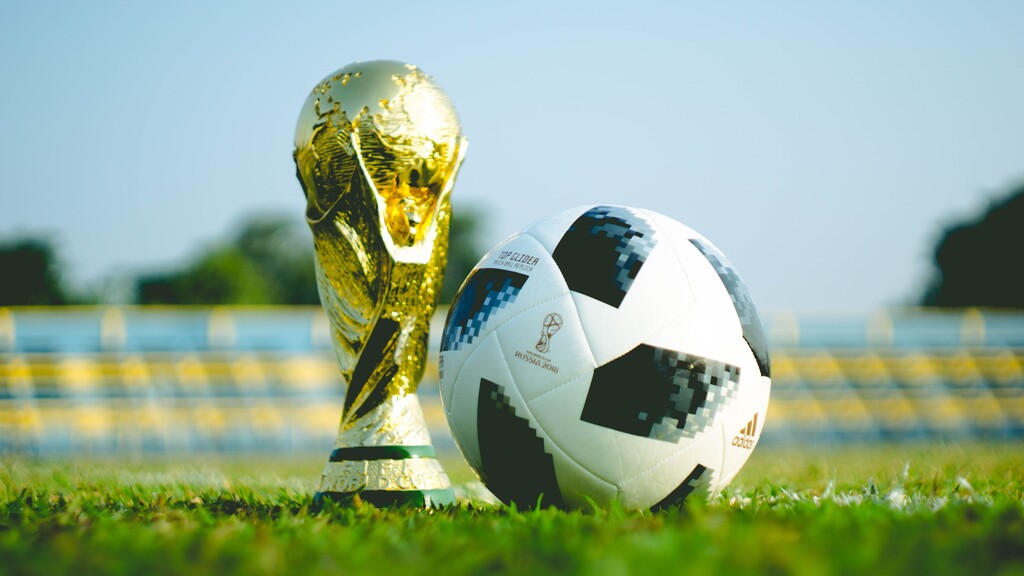 Taça da Copa do Mundo ao lado de uma bola de futebol, na grama de um campo com a arquibancada de fundo.