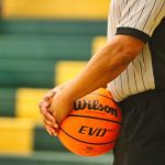 Arbitro de basquete, com camisa branca de listras pretas, segurando a bola de basquete contra o corpo.