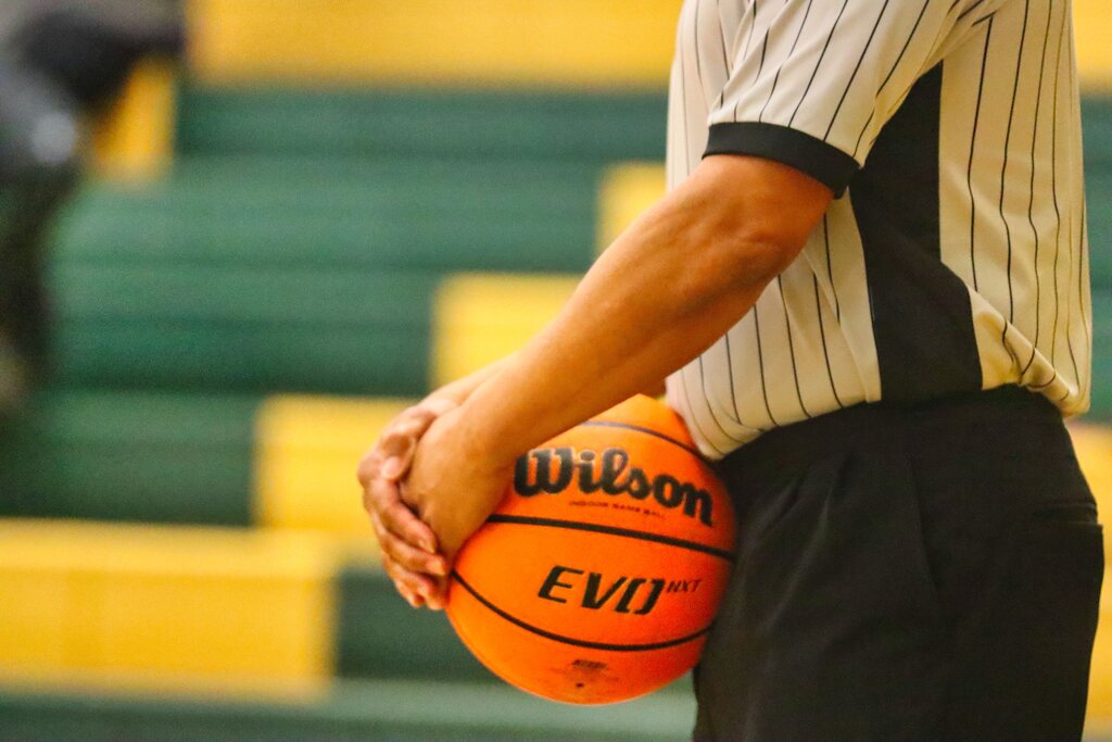 Arbitro de basquete, com camisa branca de listras pretas, segurando a bola de basquete contra o corpo.