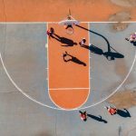 Seis pessoas jogando basquete em uma quadra cinza com um pedaço laranja. A cena é vista de cima.