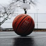 Bola de basquete laranja, escrito "NBA" em branco, deixada no chão preto da quadra, entre as linhas, de frente para a cesta.