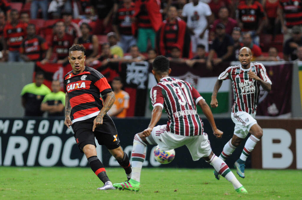 Jogador do Flamengo (vermelho e preto) encarando marcador do Fluminense (Bordô, verde e branco) chutando bola em sua direção.