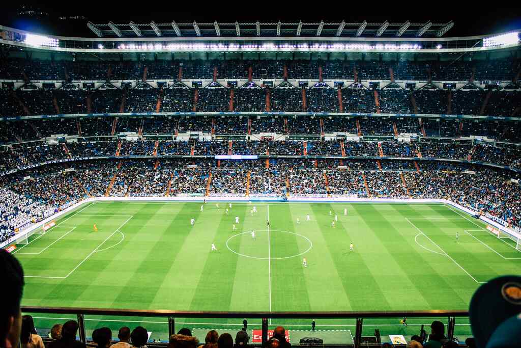 Partida de futebol vista do estádio, de uma arquibancada em nível mais elevado. Vista do resto da arquibancada do outro lado.