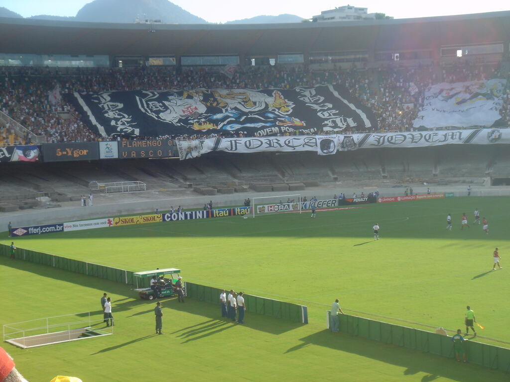Torcida do Vasco da Gama na arquibancada de cima do estádio, estendendo uma bandeira gigante com o símbolo e mascote do time.