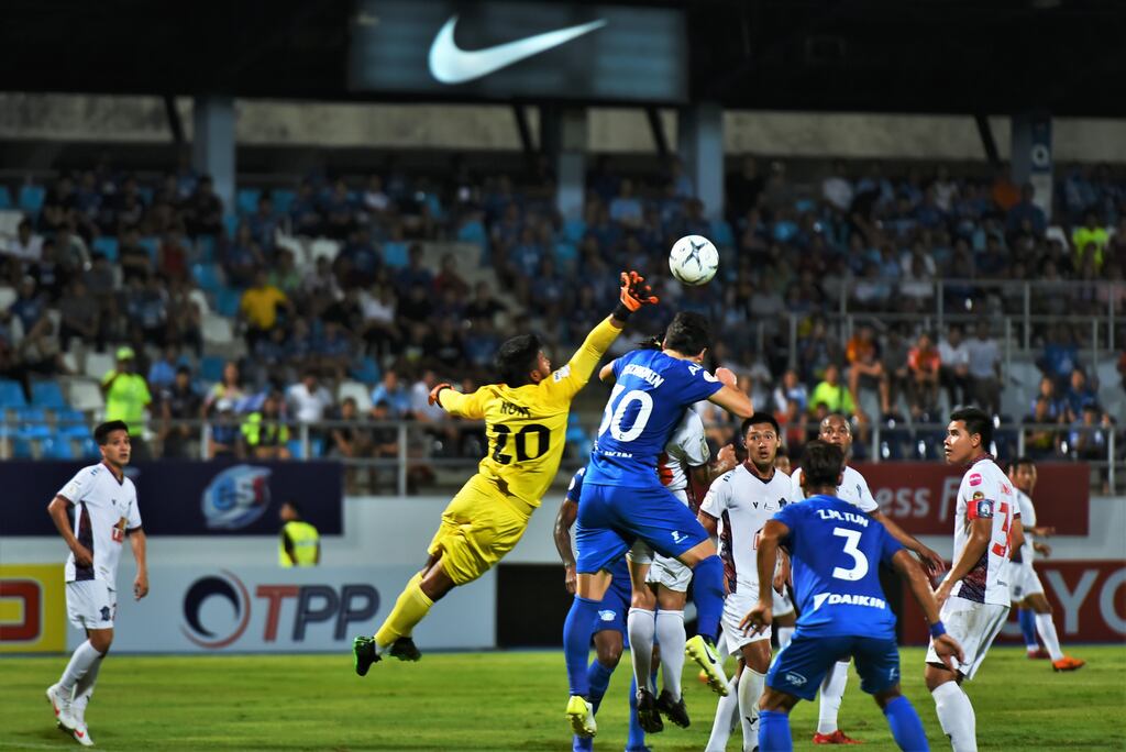 Goleiro (amarelo) saindo do gol e socando uma bola que ia em direção a cabeça do atacante (azul) durante partida de futebol.