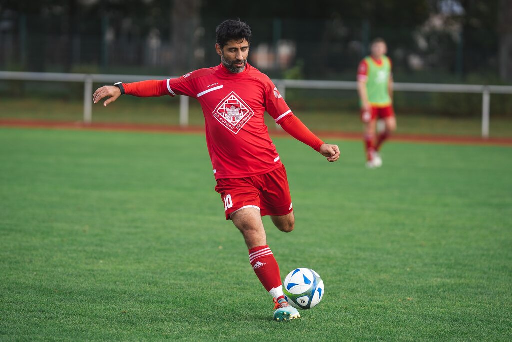 Jogador com uniforme vermelho, no campo de futebol, chutando a bola com a perna esquerda.