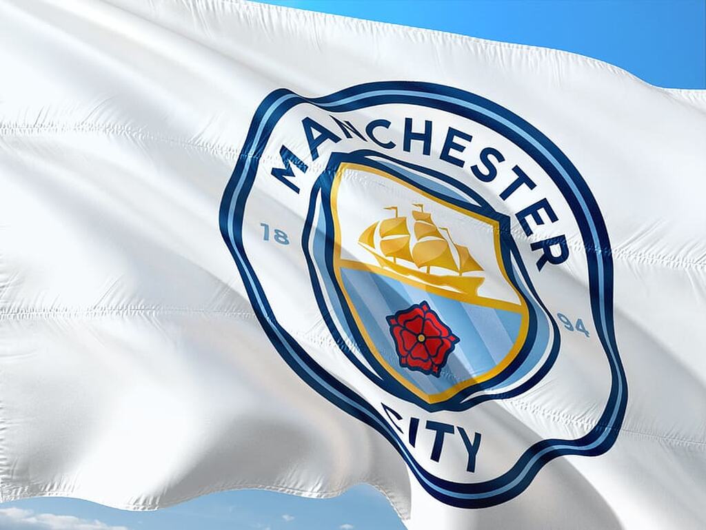 Bandeira branca com o símbolo do time Manchester City no meio e um céu azul ao fundo.
