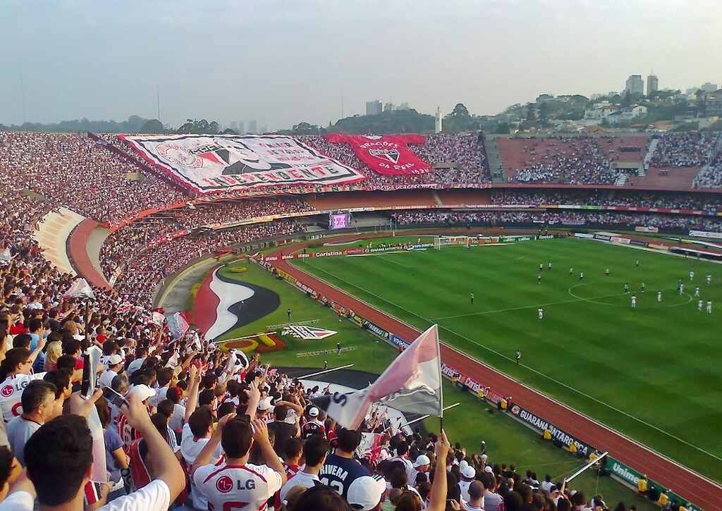 Estádio Morumbi quase cheio, com a torcida do São Pulo em festa, elevando uma bandeira e uma camisa gigante do time.
