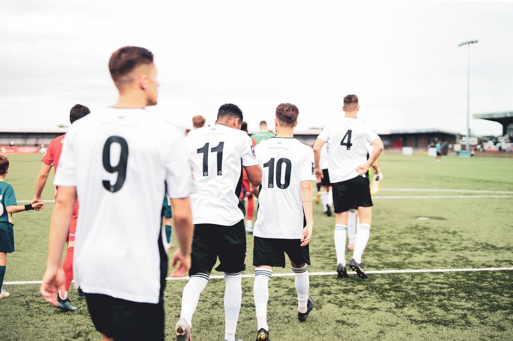 Jogadores de branco entrando em fila no campo de futebol, ao encontro dos jogadores de vermelho.