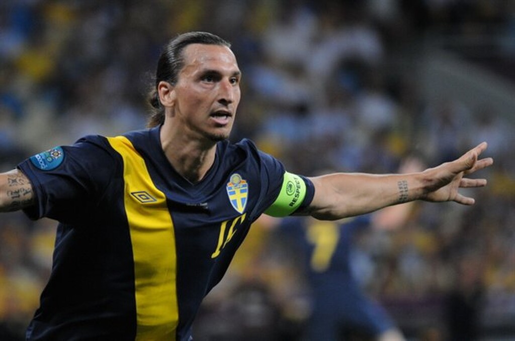 Zlatan Ibrahimovic de braços abertos, coma braçadeira de capitão, usando o uniforme azul com amarelo da seleção sueca.