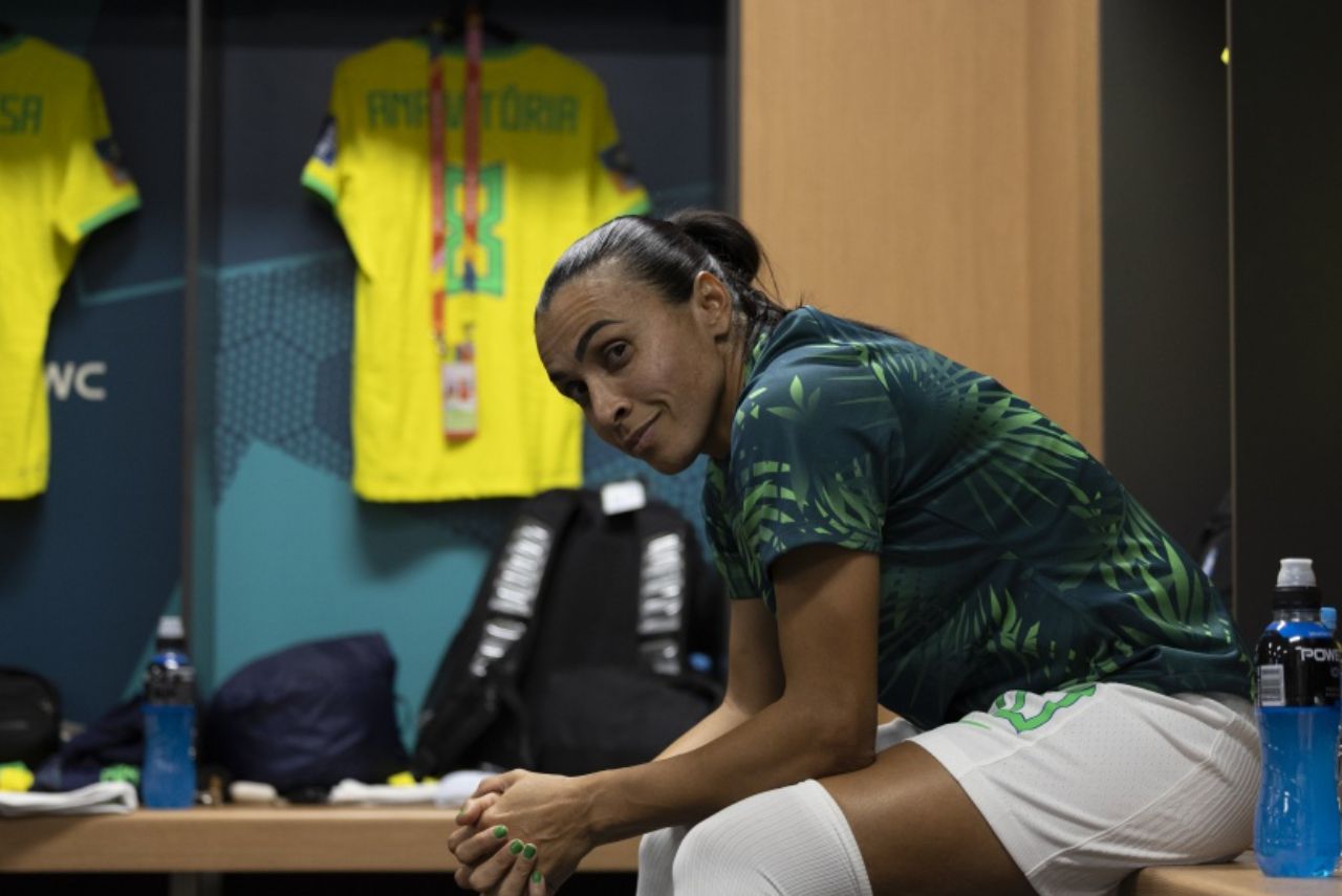 Marta, com a camisa verde da seleção, está sentada em um banco do vestiário, com os cotovelos apoiados no joelho, enquanto olha para a câmera