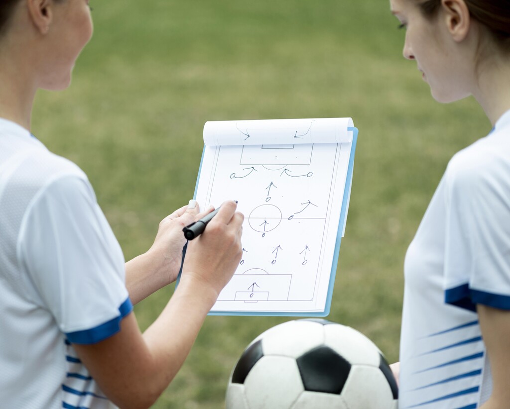 Duas garotas com roupa de futebol segurando uma bola e olhando a formação tática em um caderno