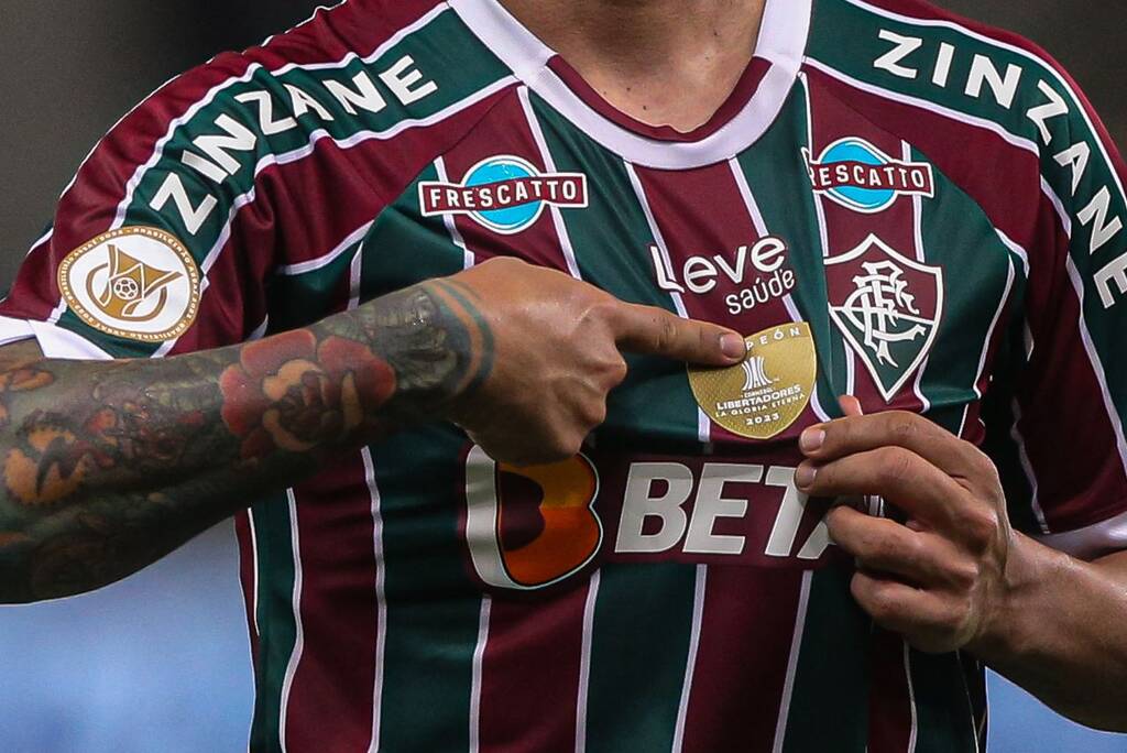 Jogador vestindo camisa do Fluminense, vermelha, verde e branca, apontando para o escudo de campeão da Libertadores.