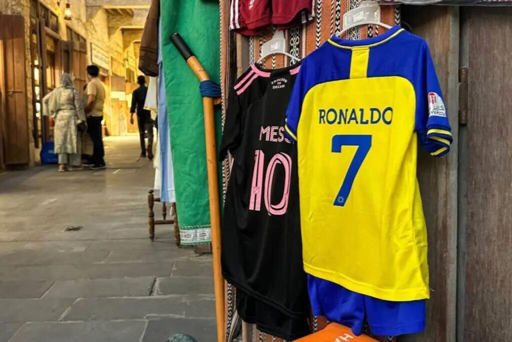 Camisa do Cristiano Ronaldo, amarela e azul, ao lado de camisa preta e rosa de Messi, em vestiário de futebol.