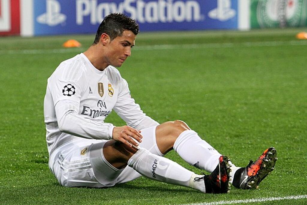 Cristiano Ronaldo, vestindo uniforme branco do Real Madrid, sentado no gramado, durante partida de futebol.