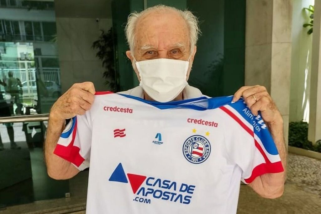 Ex-técnico de futebol Evaristo Macedo, já idoso, de máscara, segurando uma camisa branca do Bahia.