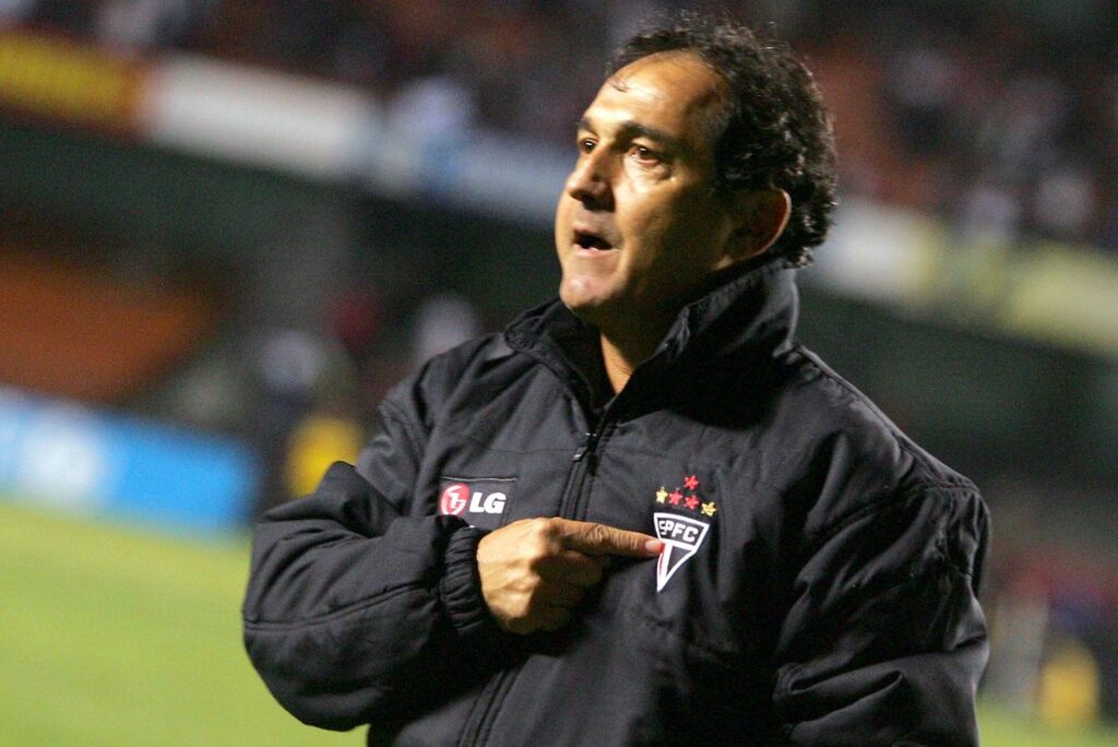 Ex-técnico de futebol Muricy Ramalho, vestindo casaco do São Paulo e apontando para escudo no peito.