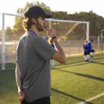 Treinador de futebol vestindo roupa cinza e boné, com apita na boca durante treino da equipe.