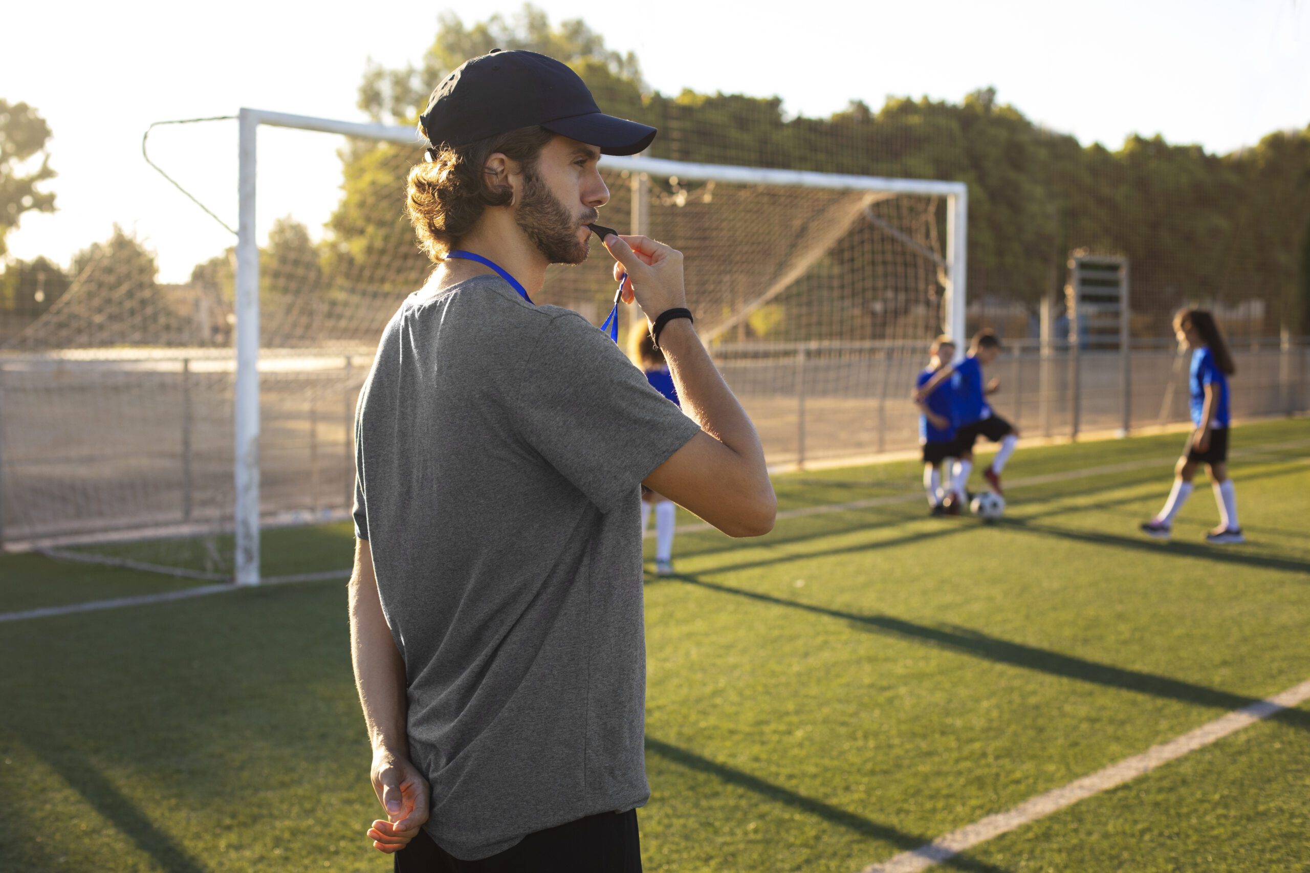 Treinador de futebol vestindo roupa cinza e boné, com apita na boca durante treino da equipe.