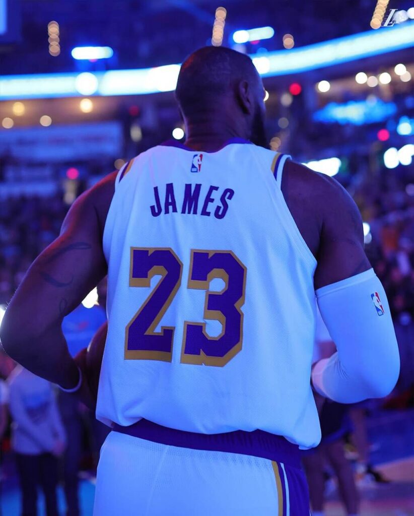 Jogador de basquete Lebron James, visto de costas com uniforme branco com detalhes azuis e amarelos, duante partida.