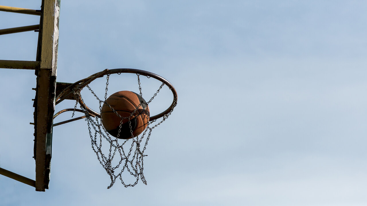 Cesta de basquete com uma bola laranja passando pelo aro e encostando na rede em um dia de sol.