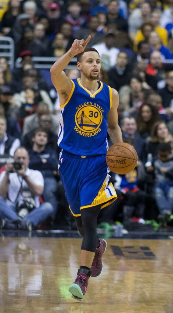 Jogador de basquete Stephen Curry, vestindo uniforme azul com detalhes amarelos e correndo durante partida.