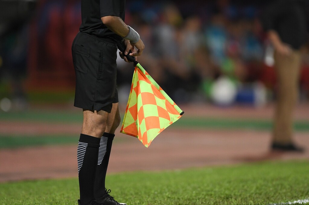 Bandeirinha de futebol vestindo uniforme preto, segurando uma bandeira laranja e amarela durante parida de futebol.