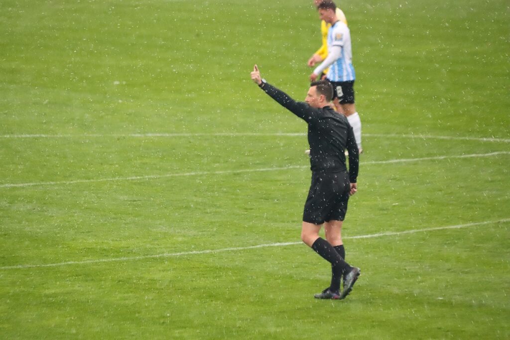 Árbitro de futebol, vestindo uniforme preto, com a mão estendida para o alto, fazendo sinal para os jogadores durante a partida.