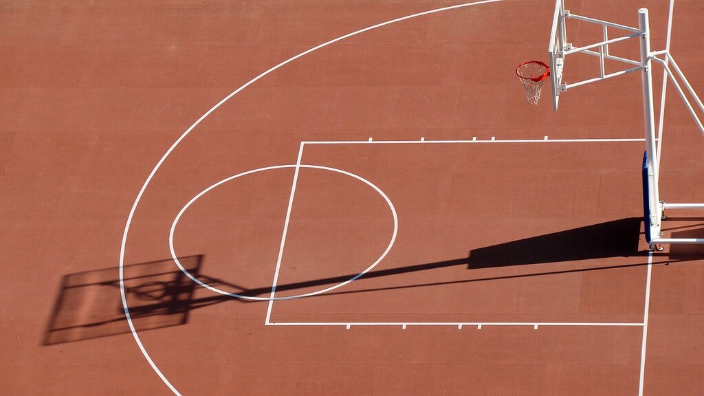 Quadra de basquete de piso marrom, com linhas brancas demarcando a área de 3 pontos, com uma cesta laranja.