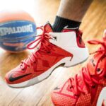 Jogador de basquete calçando um tênis vermelho com detalhes brancos, usando meia preta, com uma bola ao fundo.