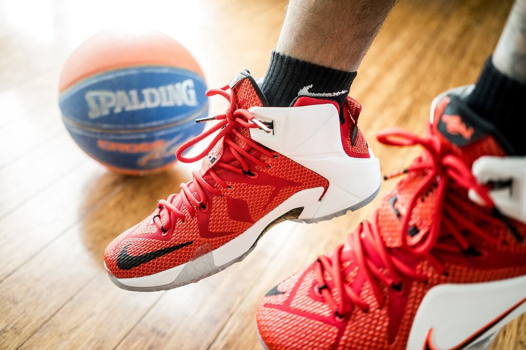 Jogador de basquete calçando um tênis vermelho com detalhes brancos, usando meia preta, com uma bola ao fundo.