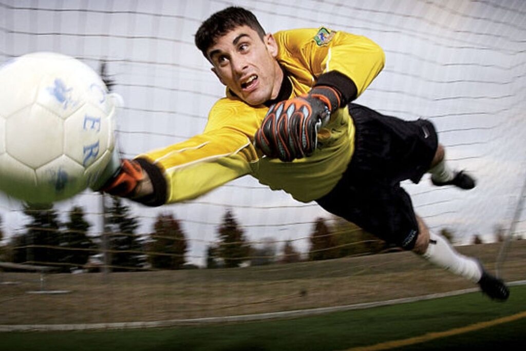 Goleiro de futebol vestindo uniforme amarelo com detalhes brancos e calça preta, defendendo um chute no gol.